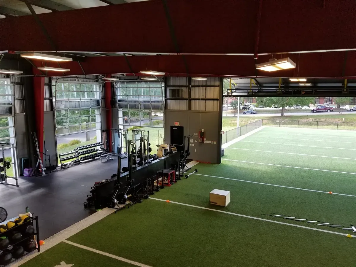 Inside an indoor training facility: strength equipment along one wall and an artificial turf pitch with ladders and hurdles laid out