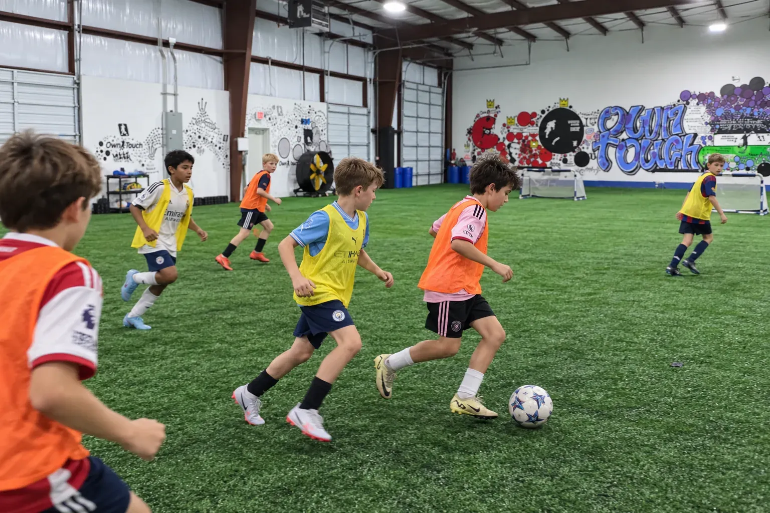 Boys in orange and yellow bibs play a small-sided match on the indoor turf at Own Touch