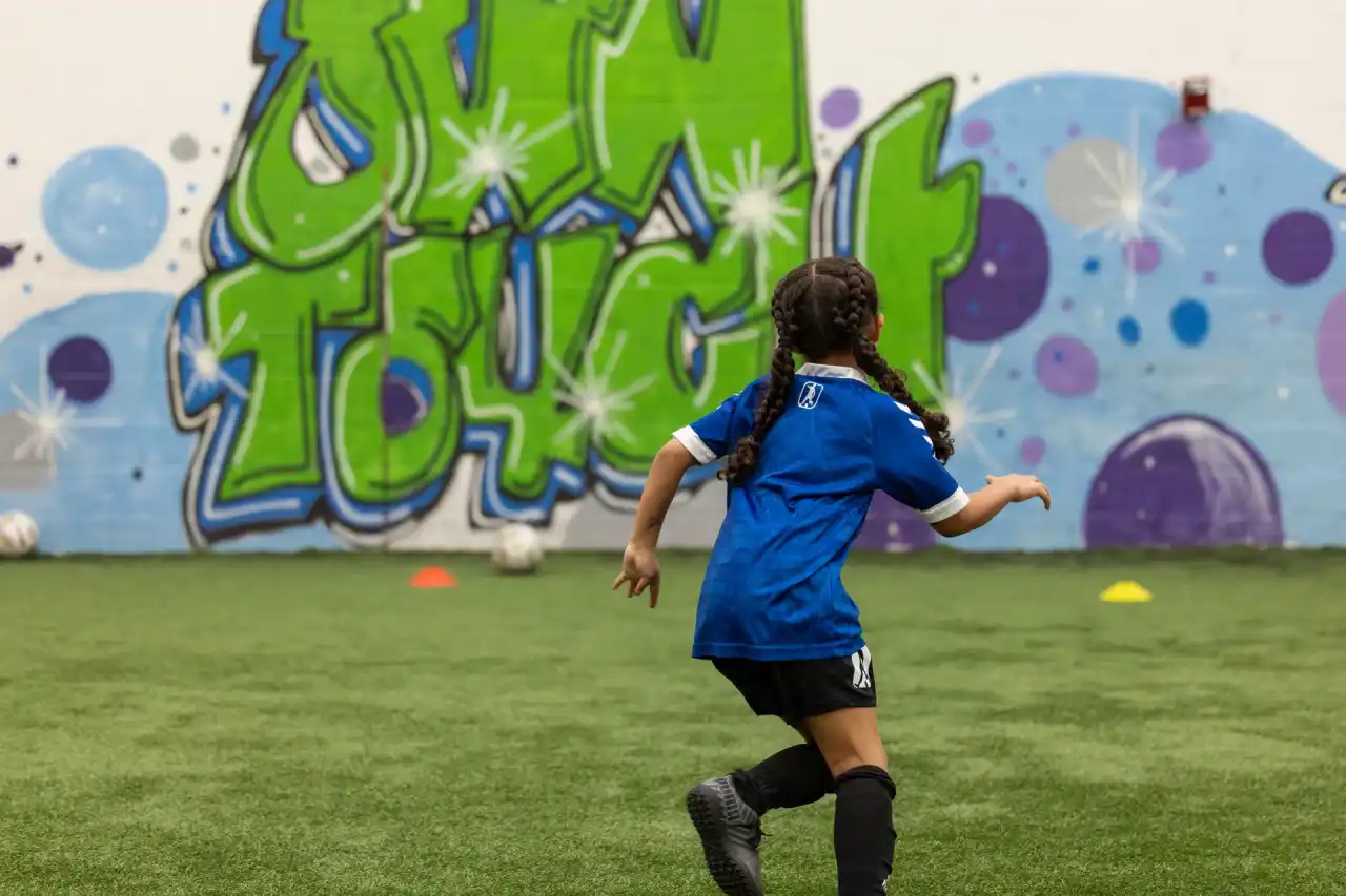 A young player in a blue kit dribbling toward the Own Touch mural on the indoor pitch
