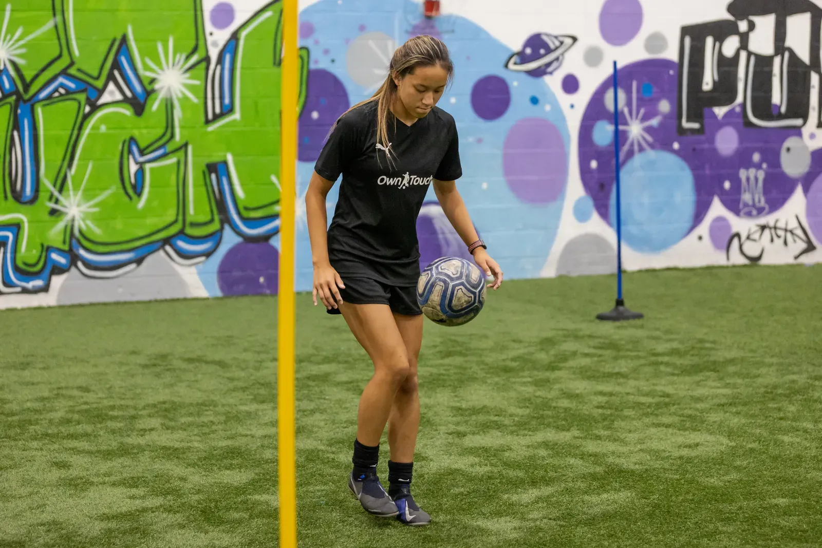 A player in a black Own Touch shirt juggling the ball between training poles
