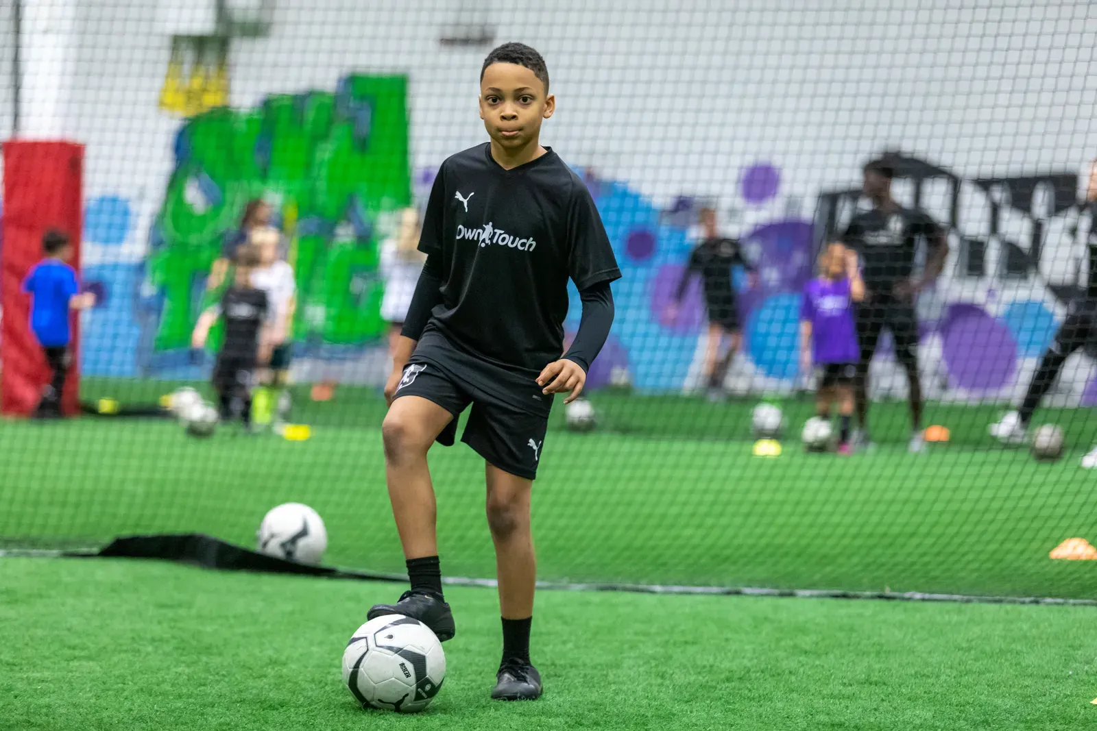 A player in a black Own Touch kit with a foot on the ball as others train behind
