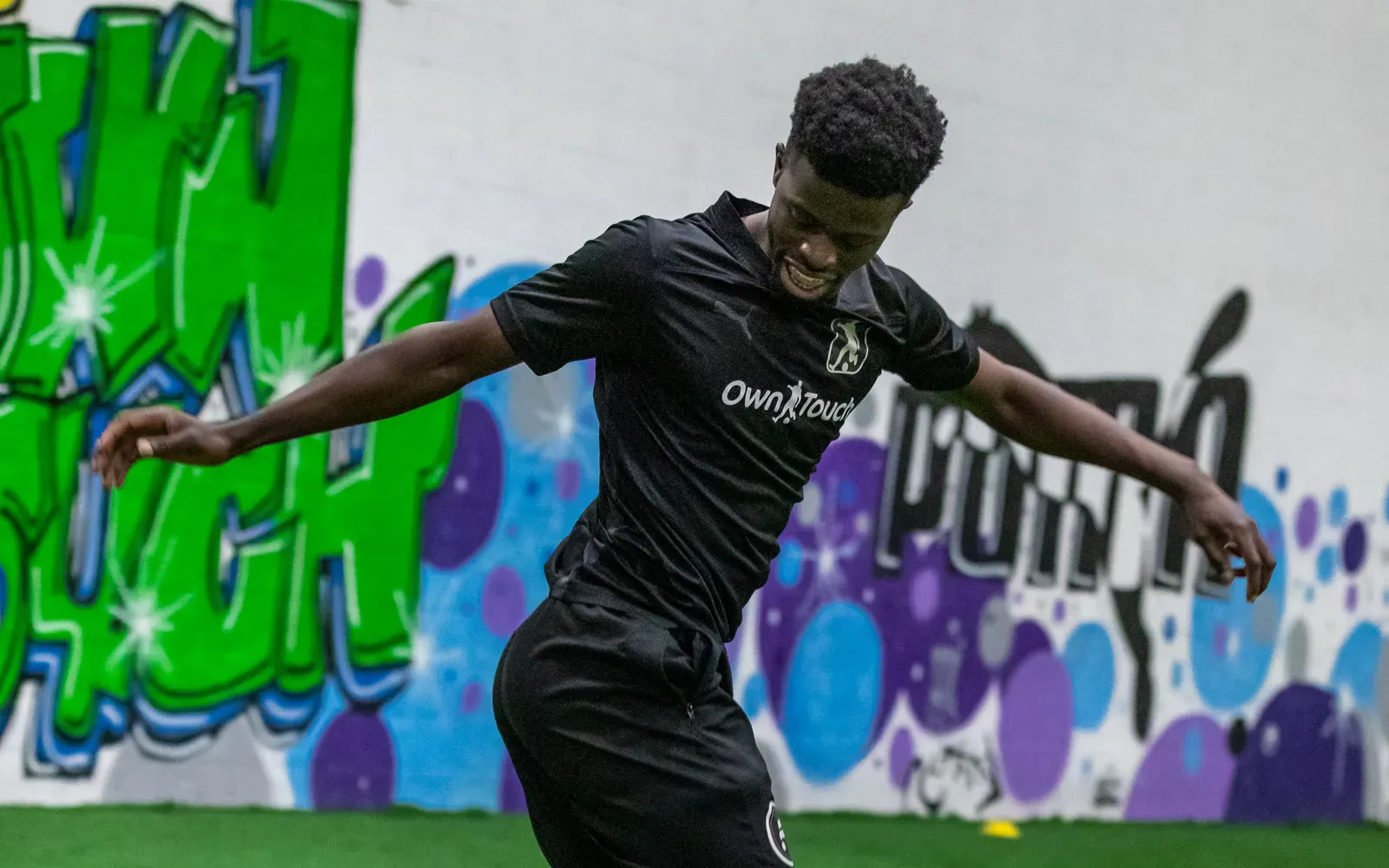 A player in a black Own Touch kit striking a volley in front of the studio mural