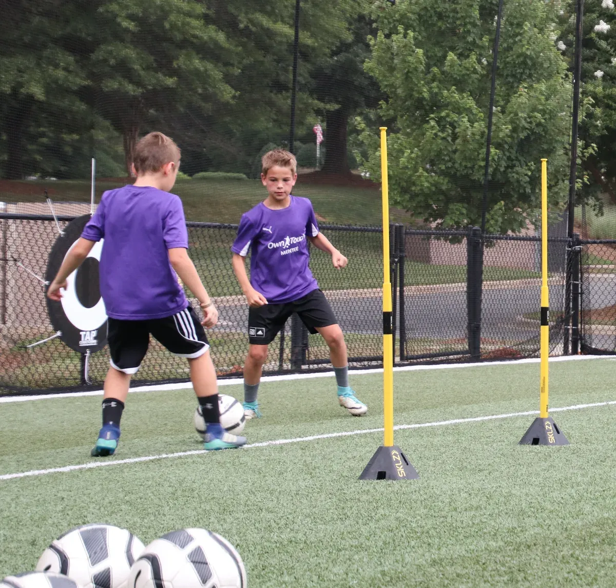 Two young players in purple Own Touch Mentee shirts working a ball through a slalom pole drill on outdoor turf
