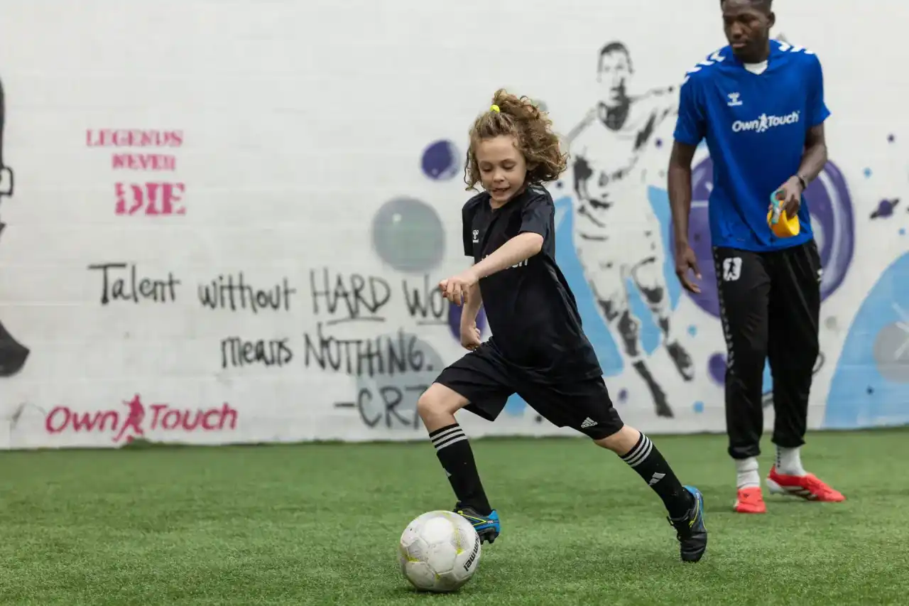 A young player dribbling the ball across the indoor turf as an Own Touch coach looks on
